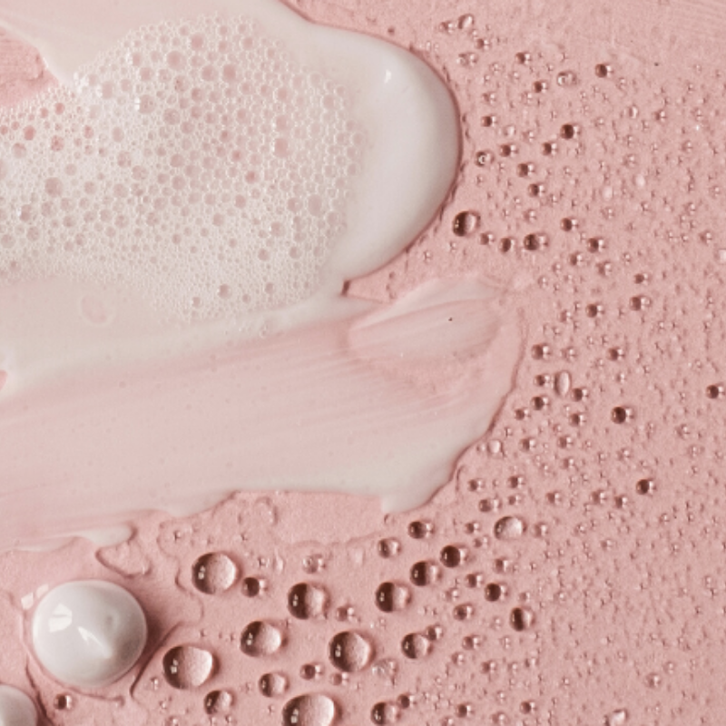 Close-up of pink skincare textures and droplets, including creamy smears, foam, and dewy water beads, representing active ingredients like AHAs in skincare