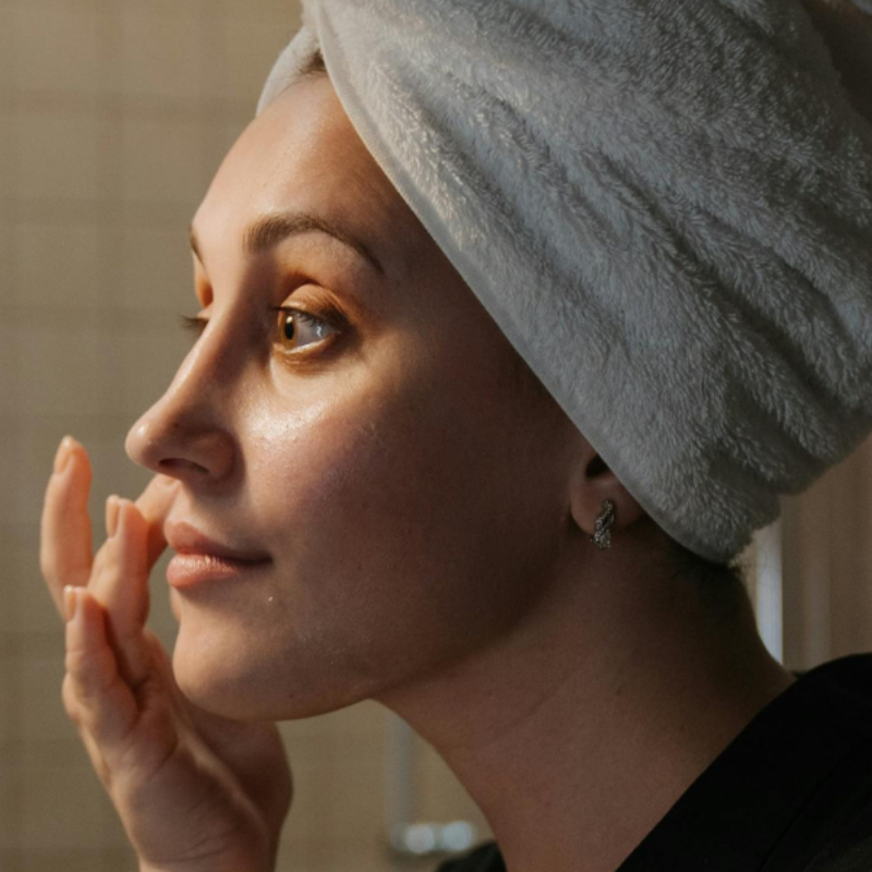 Image of woman with a towel around her head applying skincare to her face in the mirror