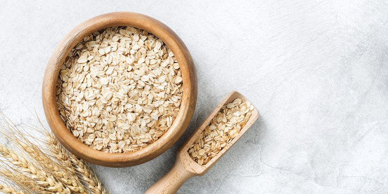 Whole oats in a wooden bowl with a matching scoop placed on a light stone background, next to wheat stalks.
