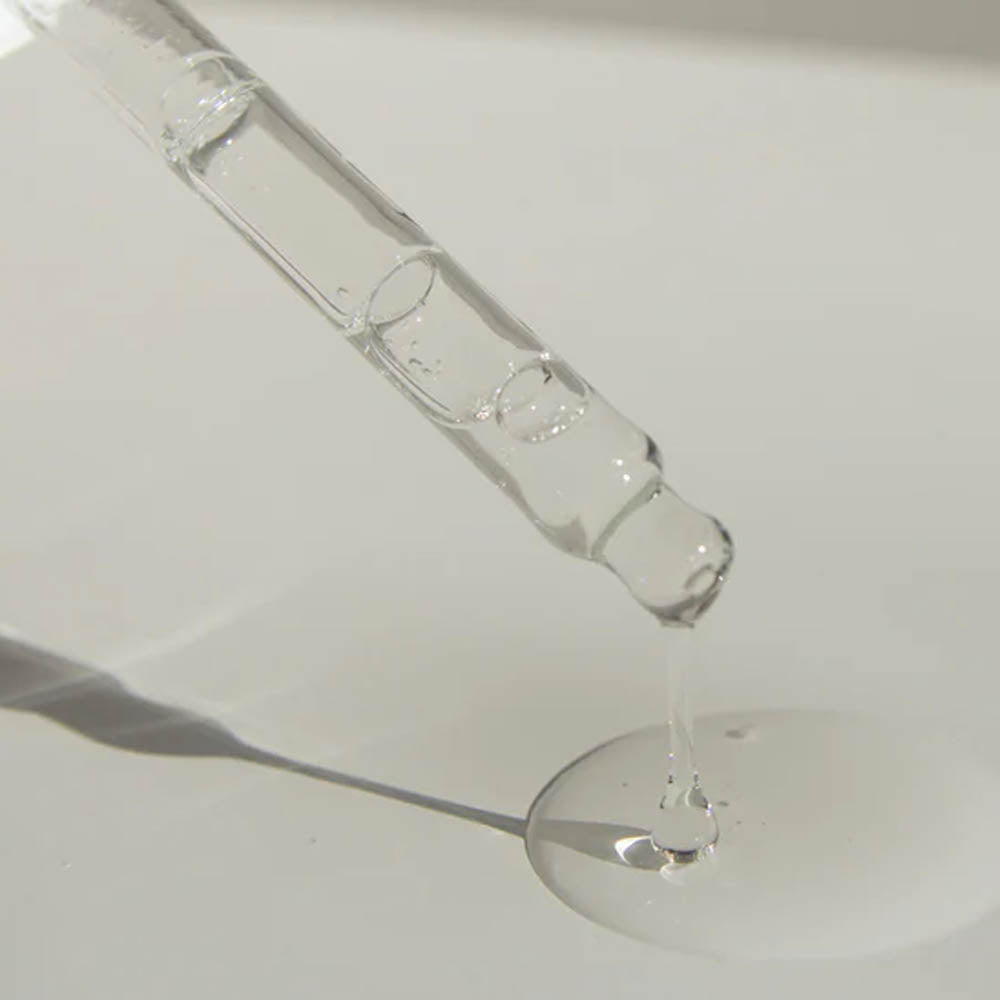 Close-up image of a glass dropper releasing a clear liquid into a pool of clear liquid symbolising a skincare serum or active ingredient like hyaluronic acid