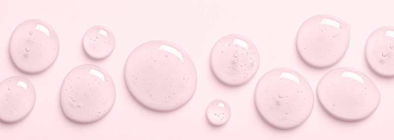 Multiple translucent pink gel-like drops of varying sizes scattered on a pale pink background, resembling a skincare serum or essence texture.