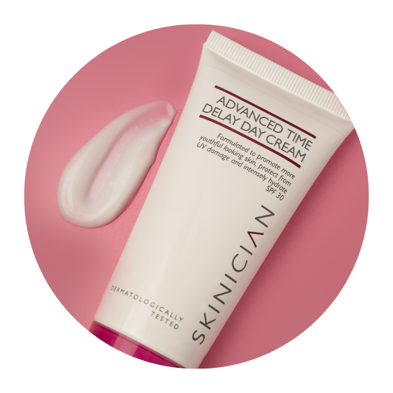 Round image of Skinician Advanced Time Delay Day Cream tube lying on a red background with the cream texture beside it