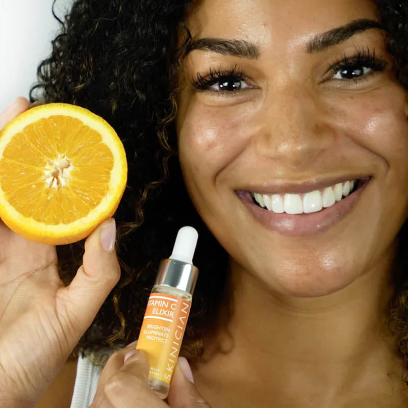 Image of a smiling female model holding a Skinician Vitamin C Elixir in one hand and a sliced orange in the other