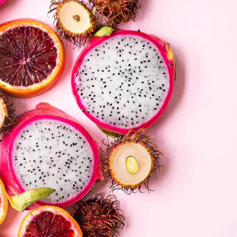 A colourful assortment of sliced tropical fruits including dragon fruit, blood orange, and rambutan on a pink background.