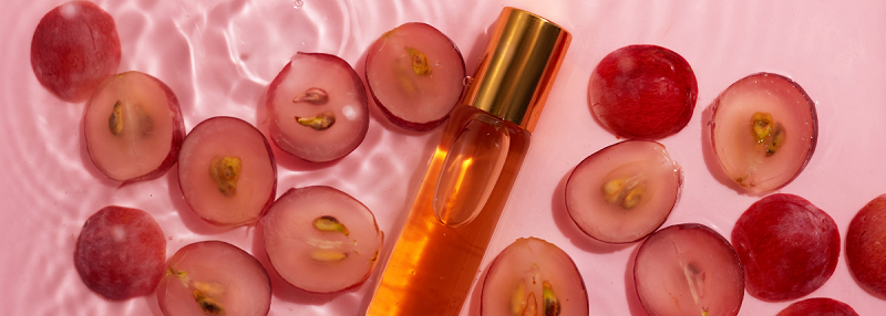 A bottle of golden grapeseed oil surrounded by halved red grapes on a rippled pink surface, symbolizing natural skincare ingredients and antioxidant-rich formulas