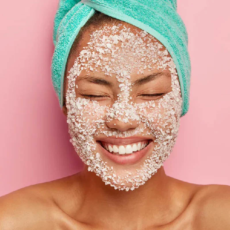 Smiling woman with eyes closed, wearing a turquoise towel on her head and a coarse facial exfoliant spread across her face.