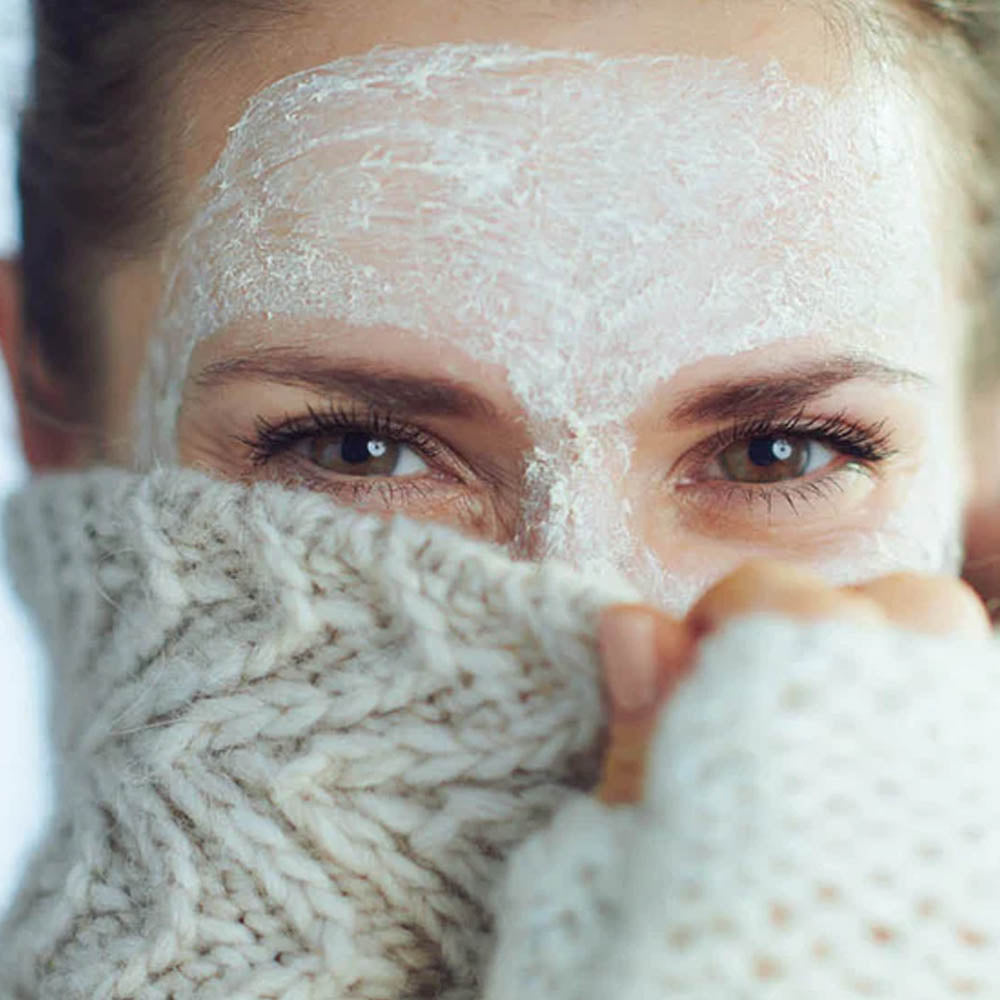 Woman with skincare face mask applied pulling a knitted jumper up to her eyes to demonstrate winter skincare