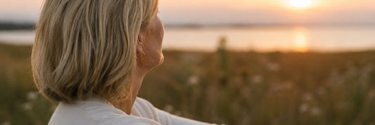 Image of an older blonde woman, looking out into the distance, with a grass landscape and sea view.