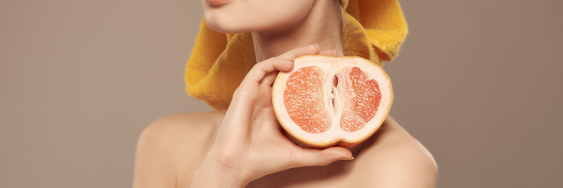 Close up image of a woman's top half, showing chin and shoulder area with her hand holding up a sliced in half grapefruit