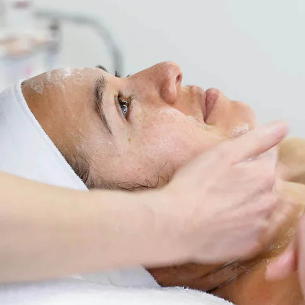 Close-up of a woman receiving a facial treatment, lying on a spa bed with a headband while a skincare product is applied to her face