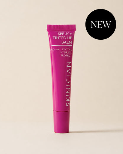 A pink tube of SKINICIAN SPF 50+ Tinted Lip Balm stands upright on a warm beige background. A black circular 'NEW' badge is in the top right corner.
