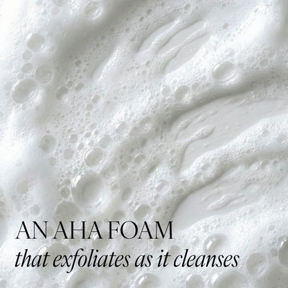 Close-up of white exfoliating foam with dense bubbles and the text 'AN AHA FOAM that exfoliates as it cleanses' overlaid at the bottom.
